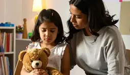 Niña en duelo abrazando un osito de peluche, acompañada por una mujer que le brinda consuelo. Escenario cálido y seguro con dibujos infantiles y luz suave, representando el apoyo emocional a niños durante la pérdida de un ser querido.